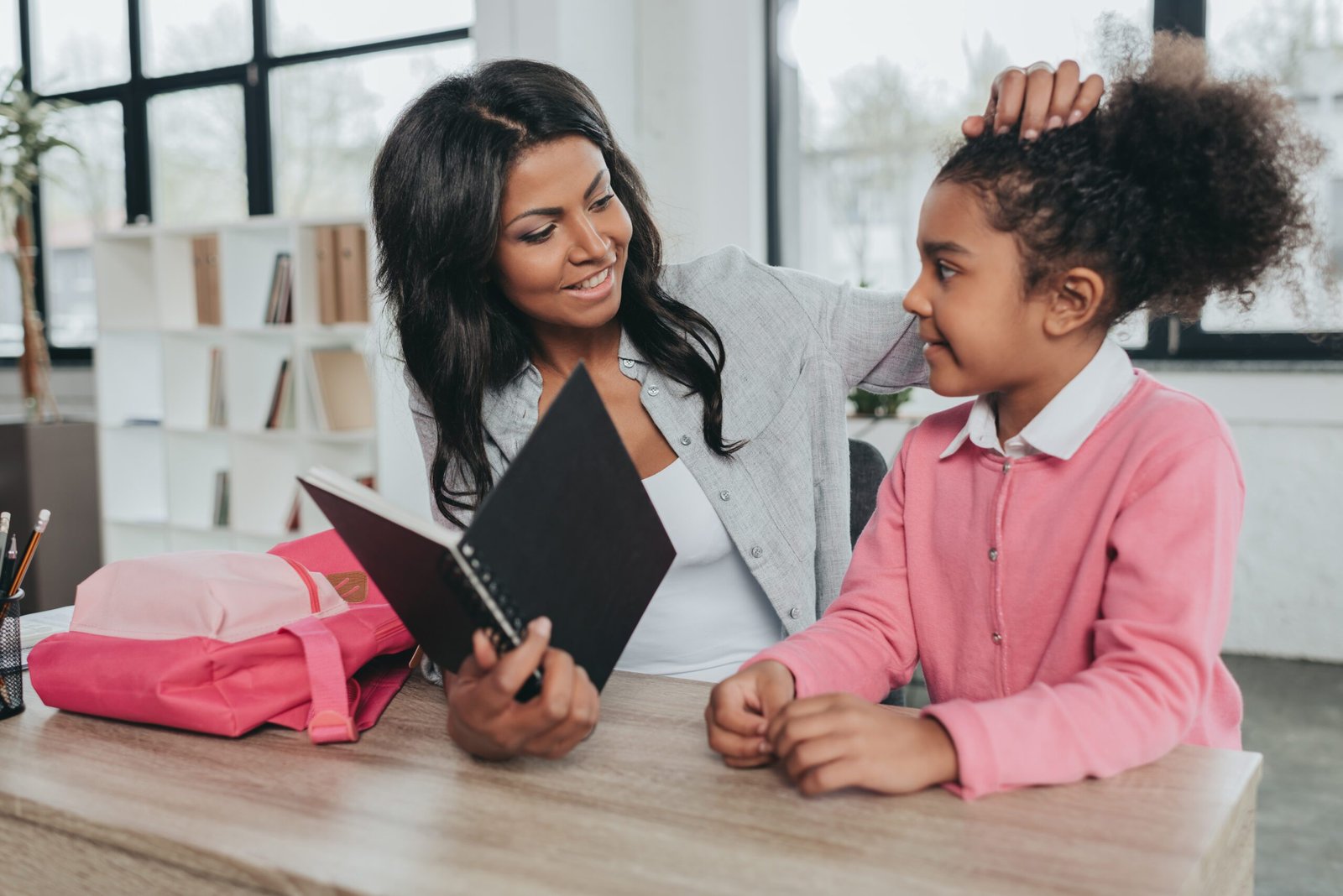 Smiling mother and daughter sitting at wooden table and reading notebook